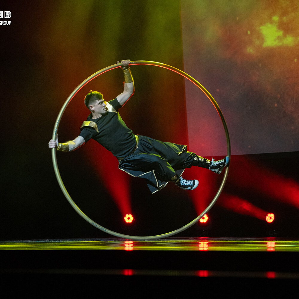 特技演員在大環（Cyr wheel）中舒展身體旋轉，紅綠燈光打亮舞台