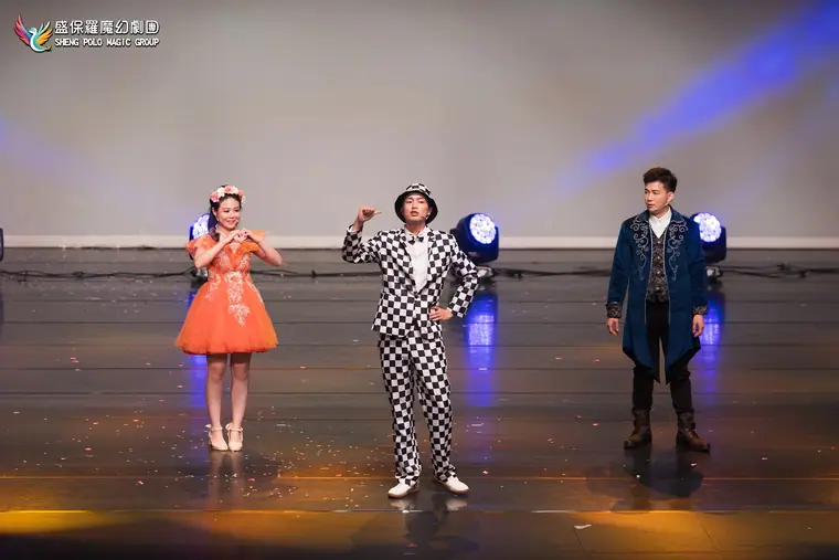 桃園展演中心展演廳《魔法師的考驗-地球危機!》 演出劇照:三位主角(橘紗裙女主、格紋西裝小丑、藍絨長袍魔法師)一字