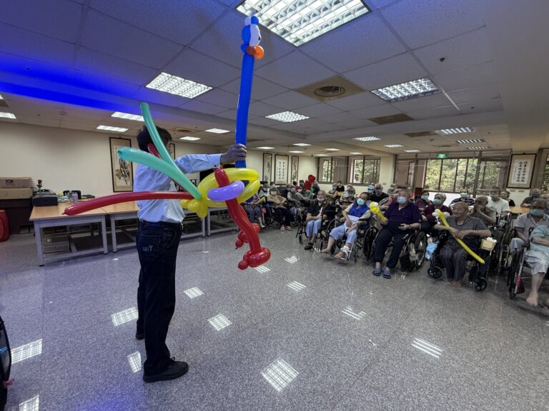 《魔法時光》桃園長照機構巡演,魔術師高舉大型氣球作品,長輩坐在台下觀看