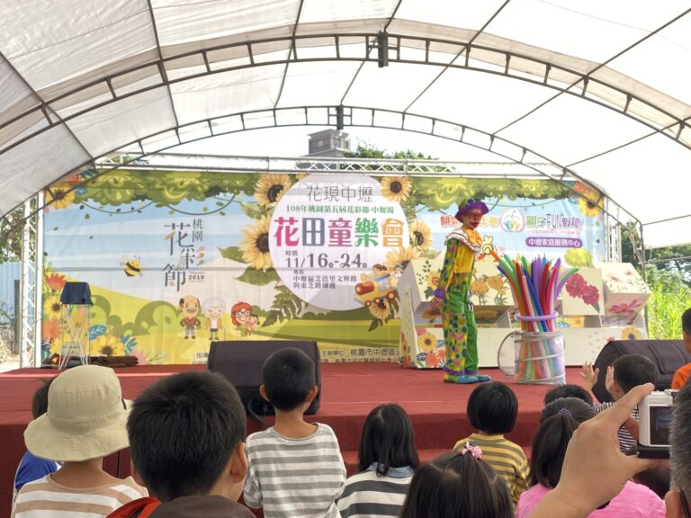 2019 桃園花彩節中壢場《小丑大氣球特技秀》，小丑在「花田童樂會」背板前表演，台下坐滿觀眾